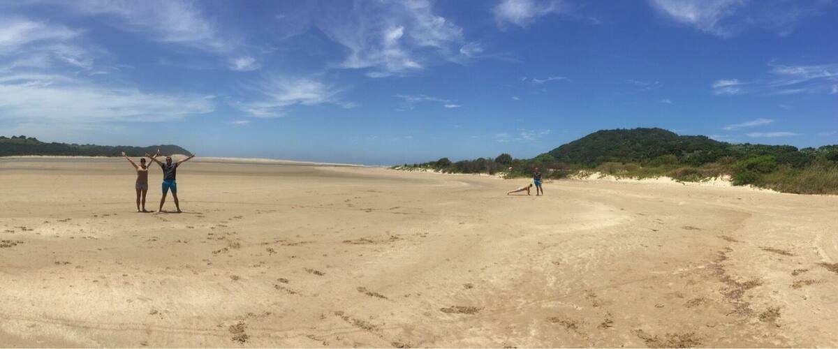 A wide sandy beach with footprints, a person lying on the sand, and hills in the background.