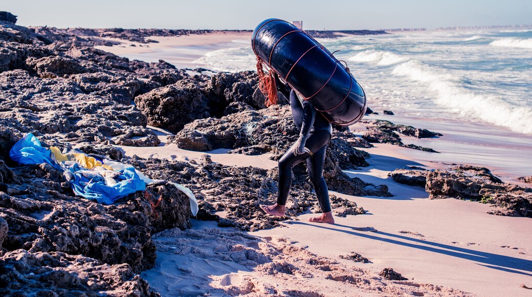 Ein felsiger Strand mit Wellen, einem Rettungsturm und einer Person, die eine Boje trägt.