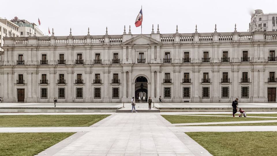 A large neoclassical building with a central entrance, surrounded by well-maintained lawns and a few people walking around.