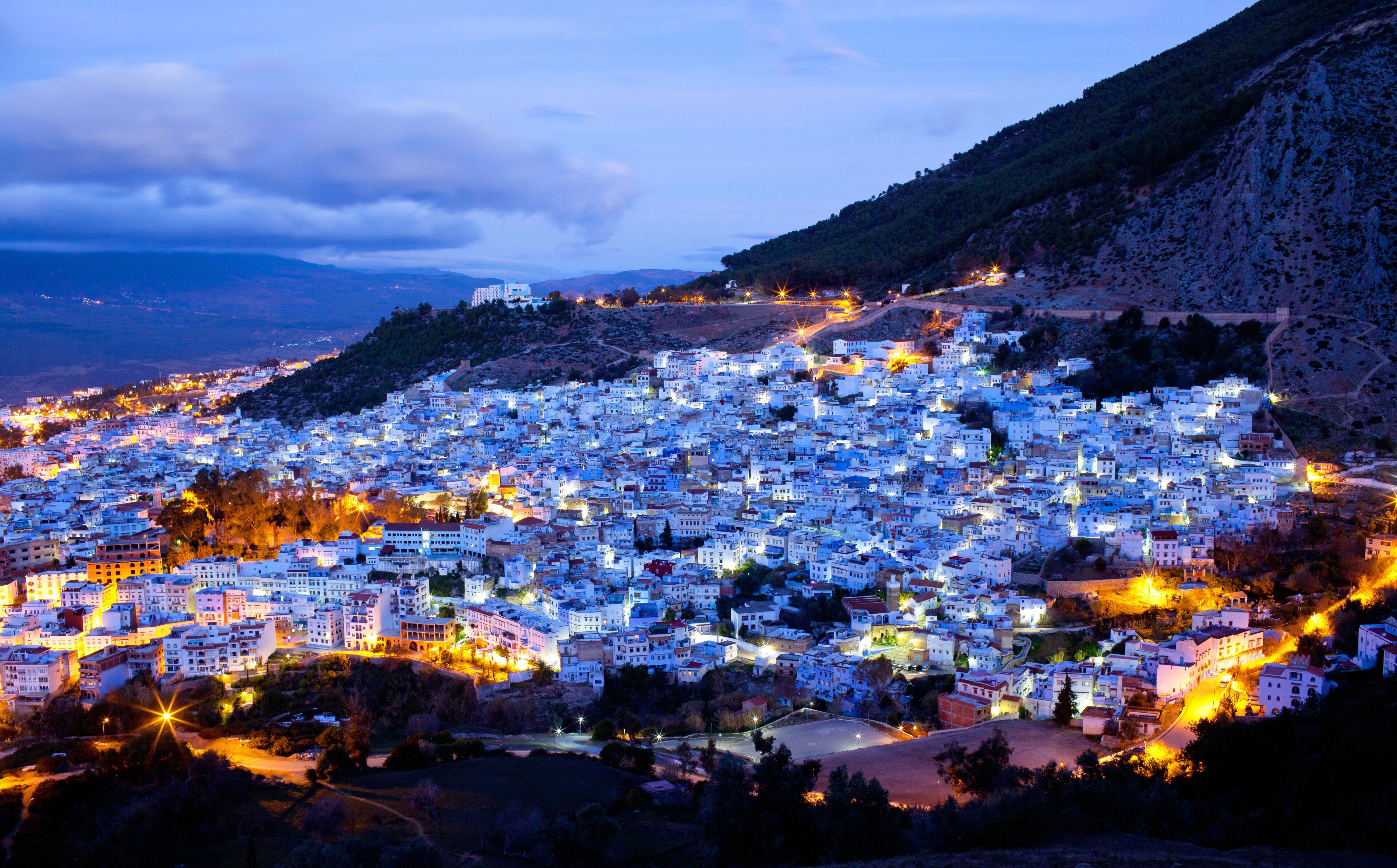 A densely packed town with white buildings, a mountain in the background, and a river in the foreground.