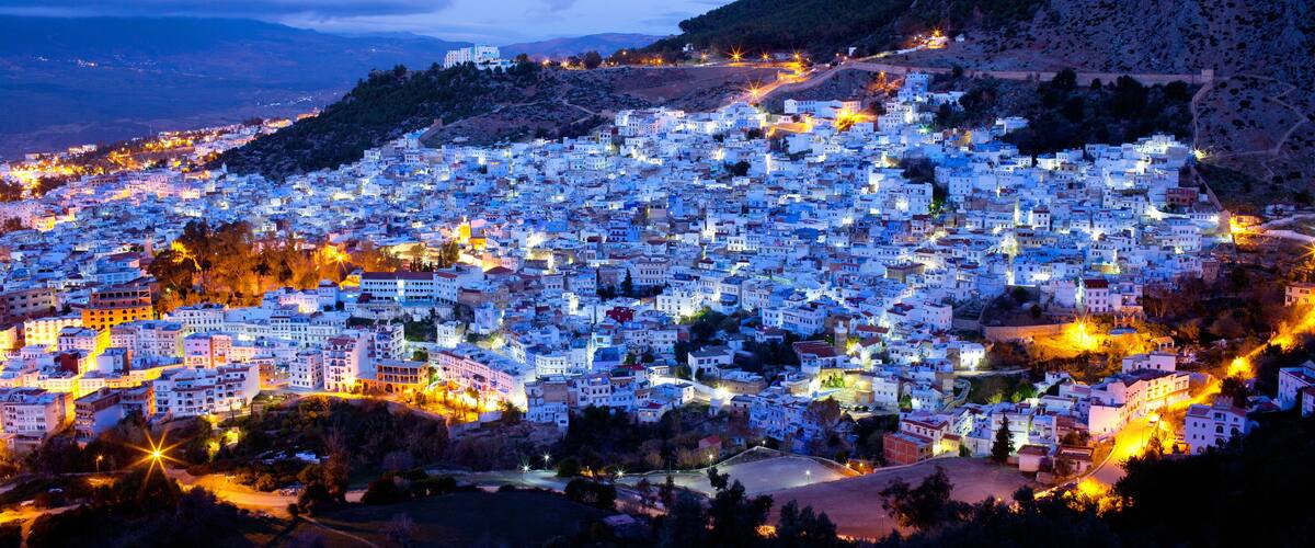 A densely packed town with white buildings, a mountain in the background, and a river in the foreground.