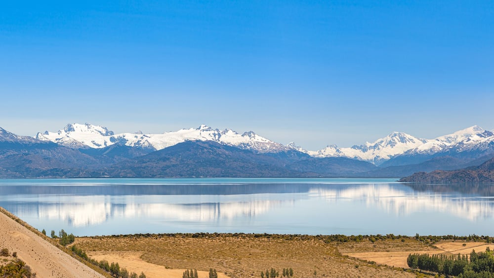 A mountain range with snow-capped peaks, a calm lake, and a barren landscape with sparse vegetation.