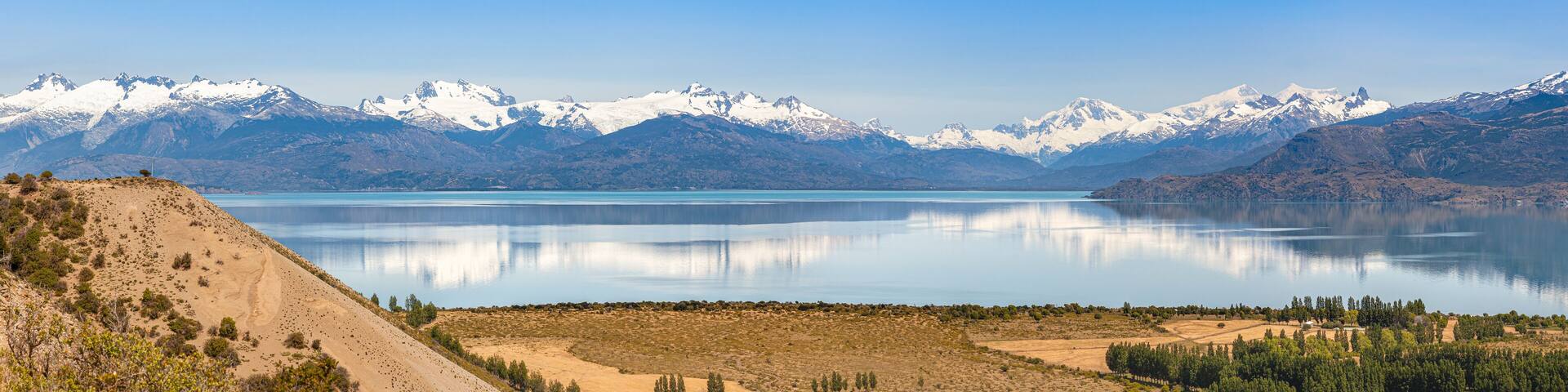 A mountain range with snow-capped peaks, a calm lake, and a barren landscape with sparse vegetation.