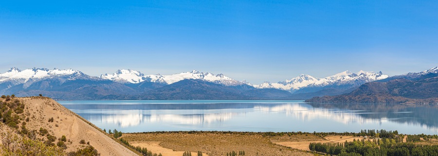 A mountain range with snow-capped peaks, a calm lake, and a barren landscape with sparse vegetation.
