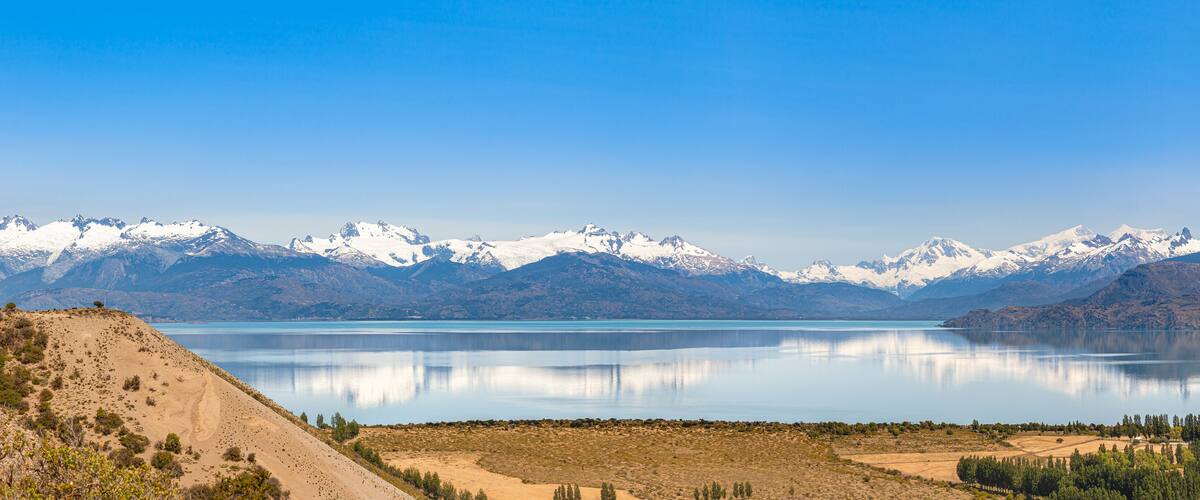 A mountain range with snow-capped peaks, a calm lake, and a barren landscape with sparse vegetation.