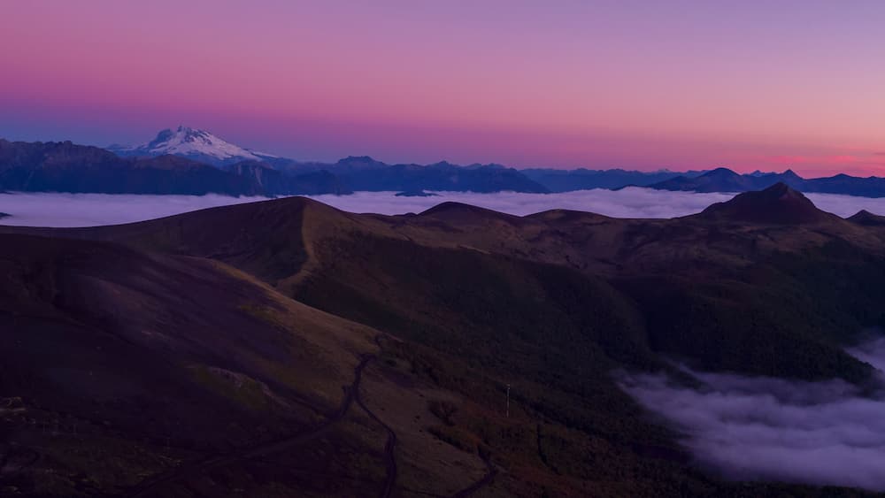 A mountain range with a snow-capped peak, valleys, and a layer of clouds below.
