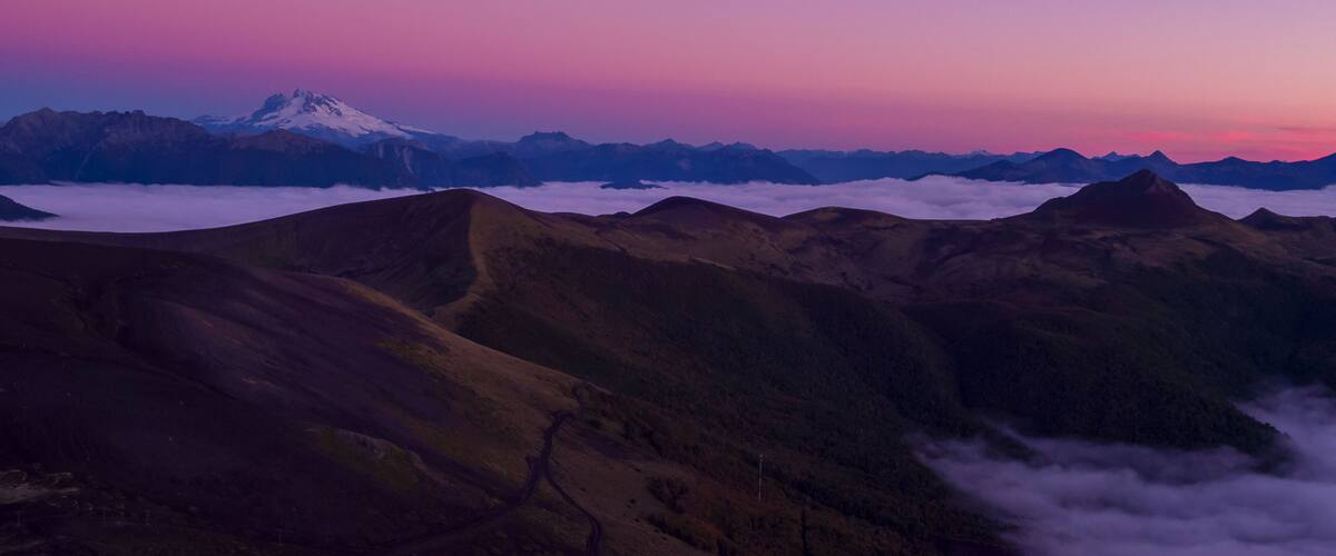 A mountain range with a snow-capped peak, valleys, and a layer of clouds below.