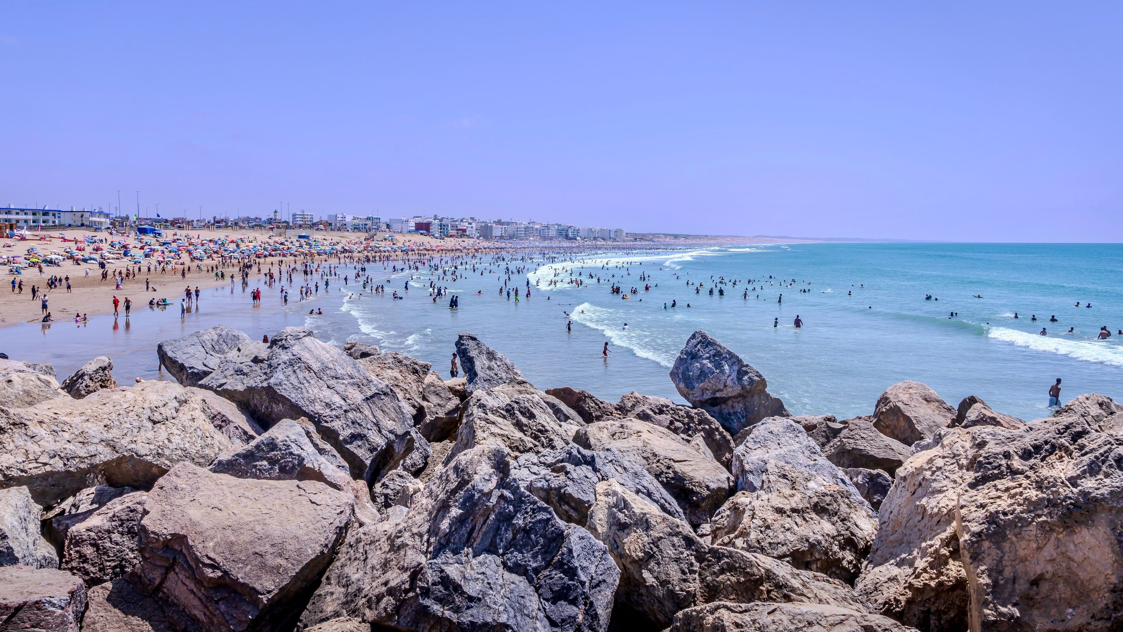 A crowded beach with rocky foreground and a clear blue sky.