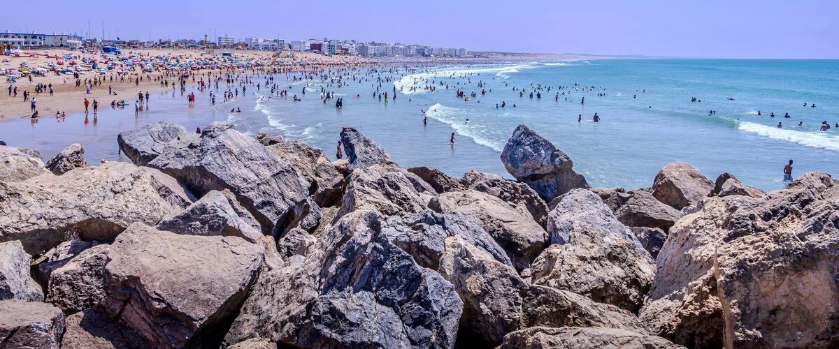 A crowded beach with rocky foreground and a clear blue sky.