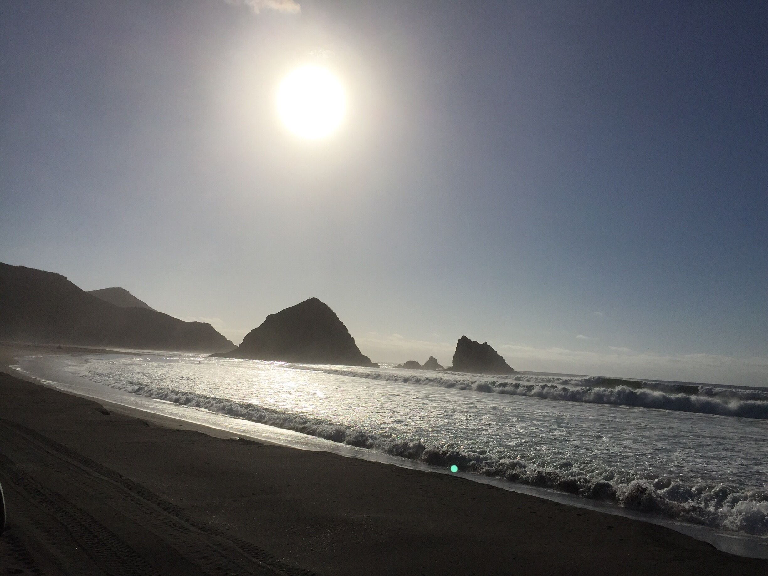 A beach with waves, a clear sky, and mountains in the background.