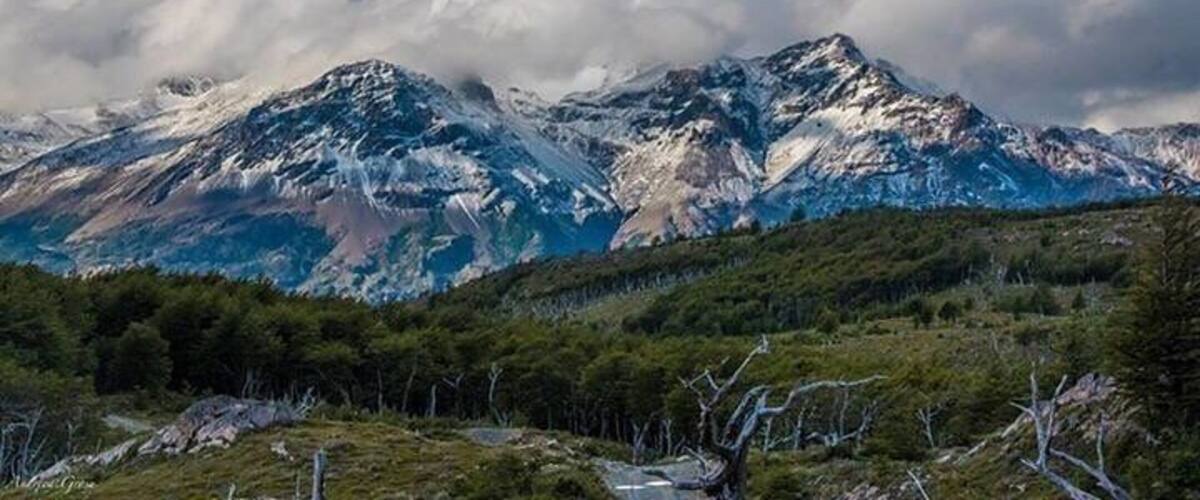 A mountain range with snow-capped peaks, a forested valley, and a cloudy sky.