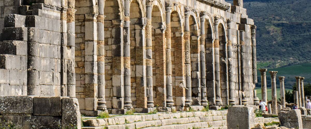 Ancient stone ruins with columns and arches, set against a mountainous backdrop.