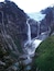 A glacier waterfall in a canyon with steep cliffs and a small lake at the base.
