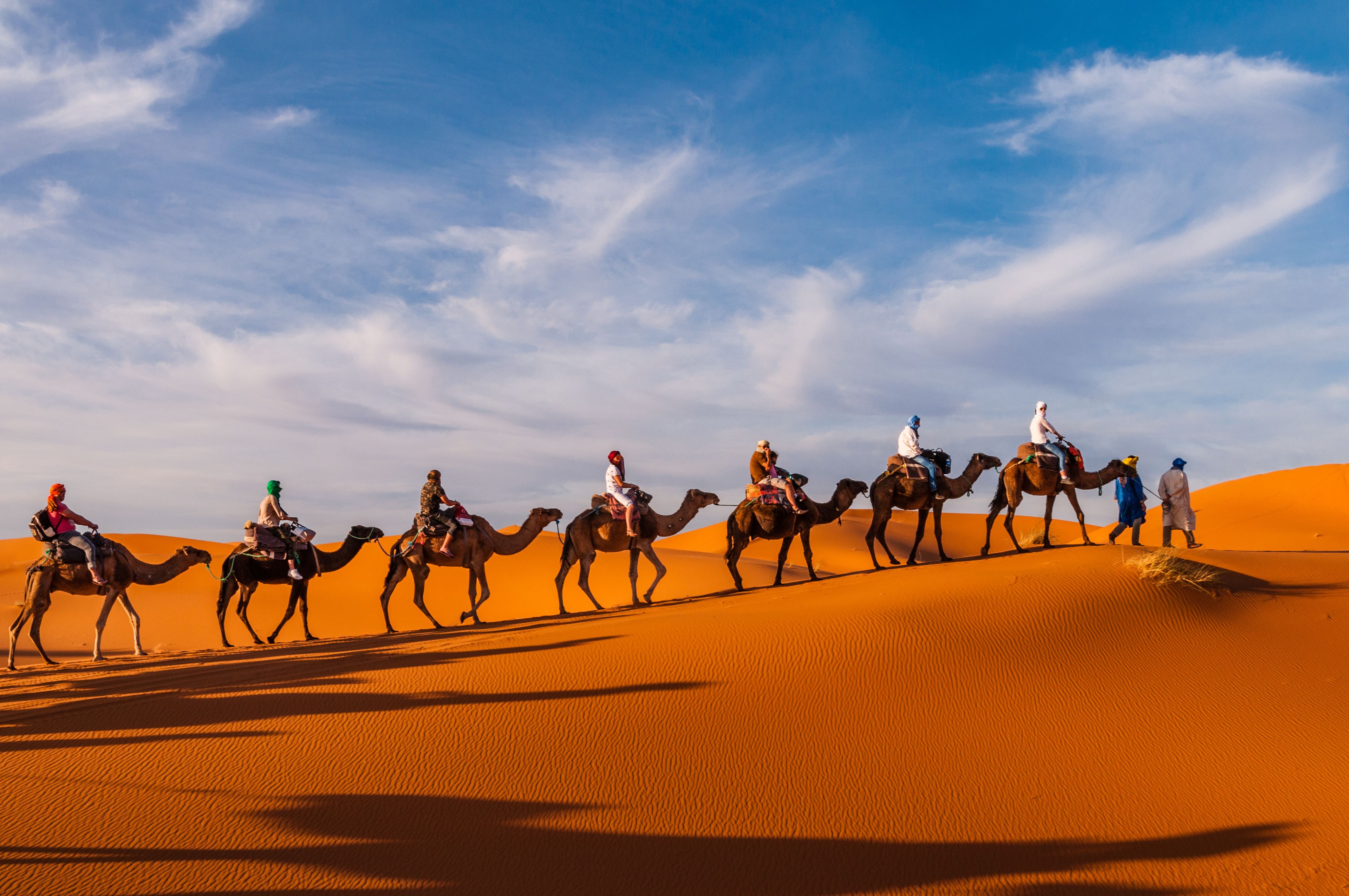 Eine Gruppe von Menschen reitet auf Kamelen durch eine Wüstenlandschaft mit Sanddünen.