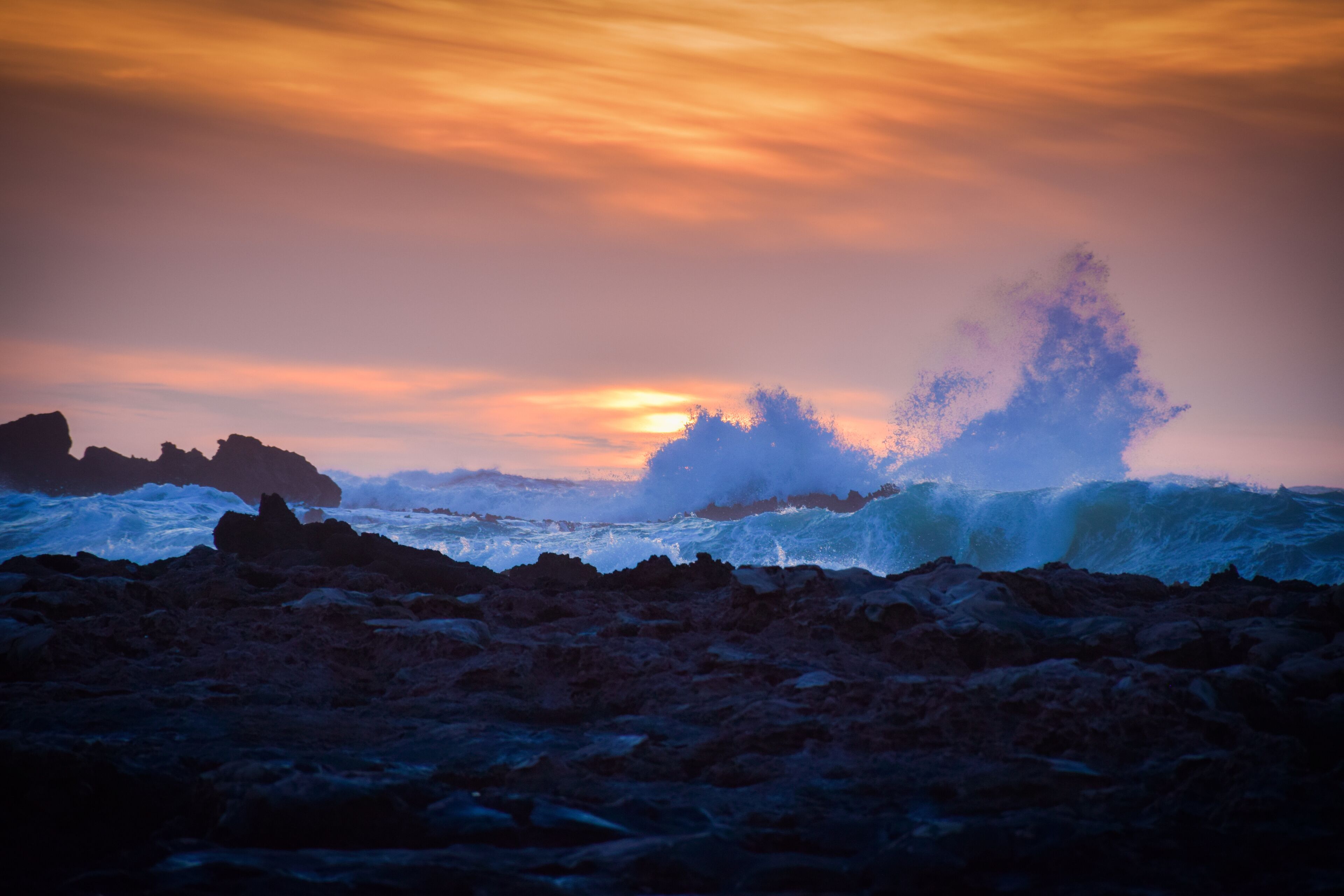 A rocky shoreline with waves crashing against the rocks under a sunset sky.