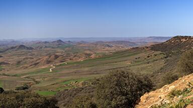 A panoramic view of rolling hills, a winding road, and distant mountains under a clear sky.