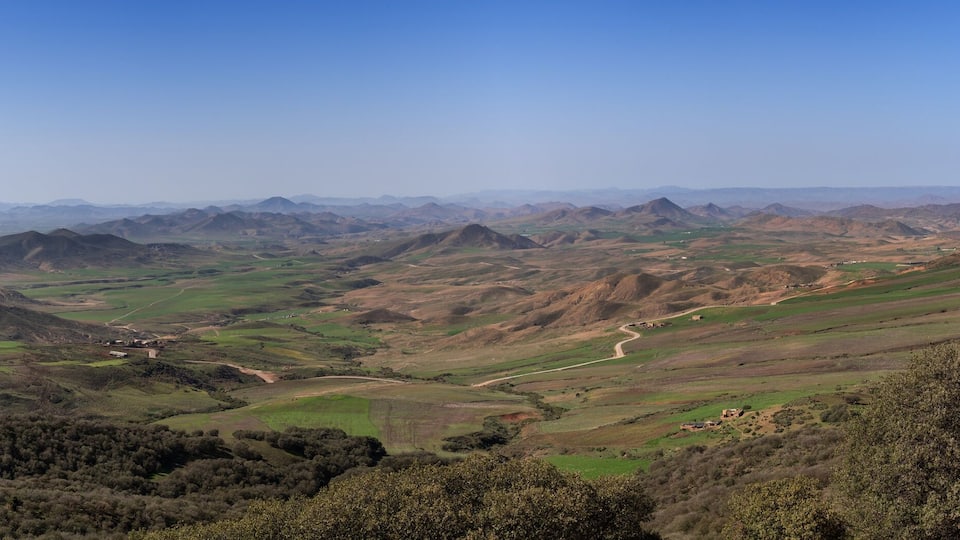 A panoramic view of rolling hills, a winding road, and distant mountains under a clear sky.