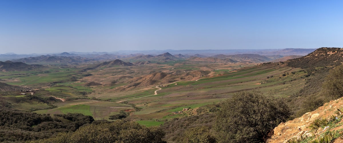 A panoramic view of rolling hills, a winding road, and distant mountains under a clear sky.