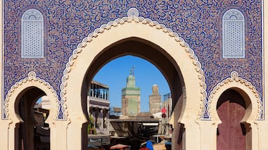 Um arco tradicional com detalhada decoração em azulejos azuis e brancos, que dá acesso a uma vista de dois minaretes ao longe.