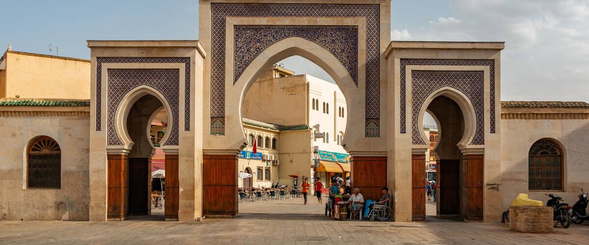 An ornate archway with intricate tile work, leading into a bustling street with cafes and pedestrians.
