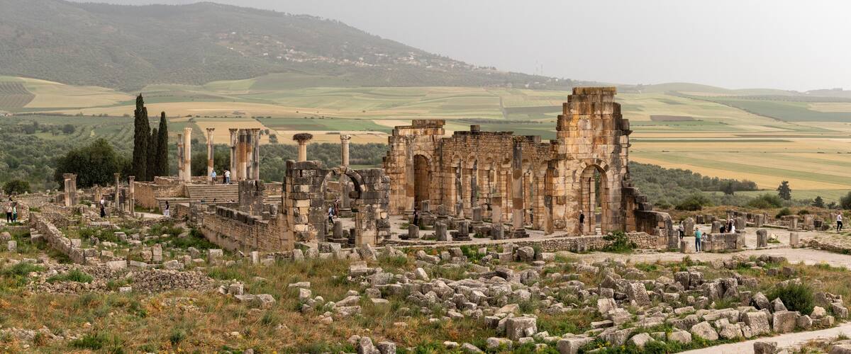Ancient ruins with columns and arches, surrounded by rolling hills and fields.