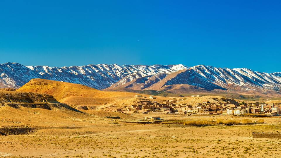 A desert landscape with a small settlement and snow-capped mountains in the background.