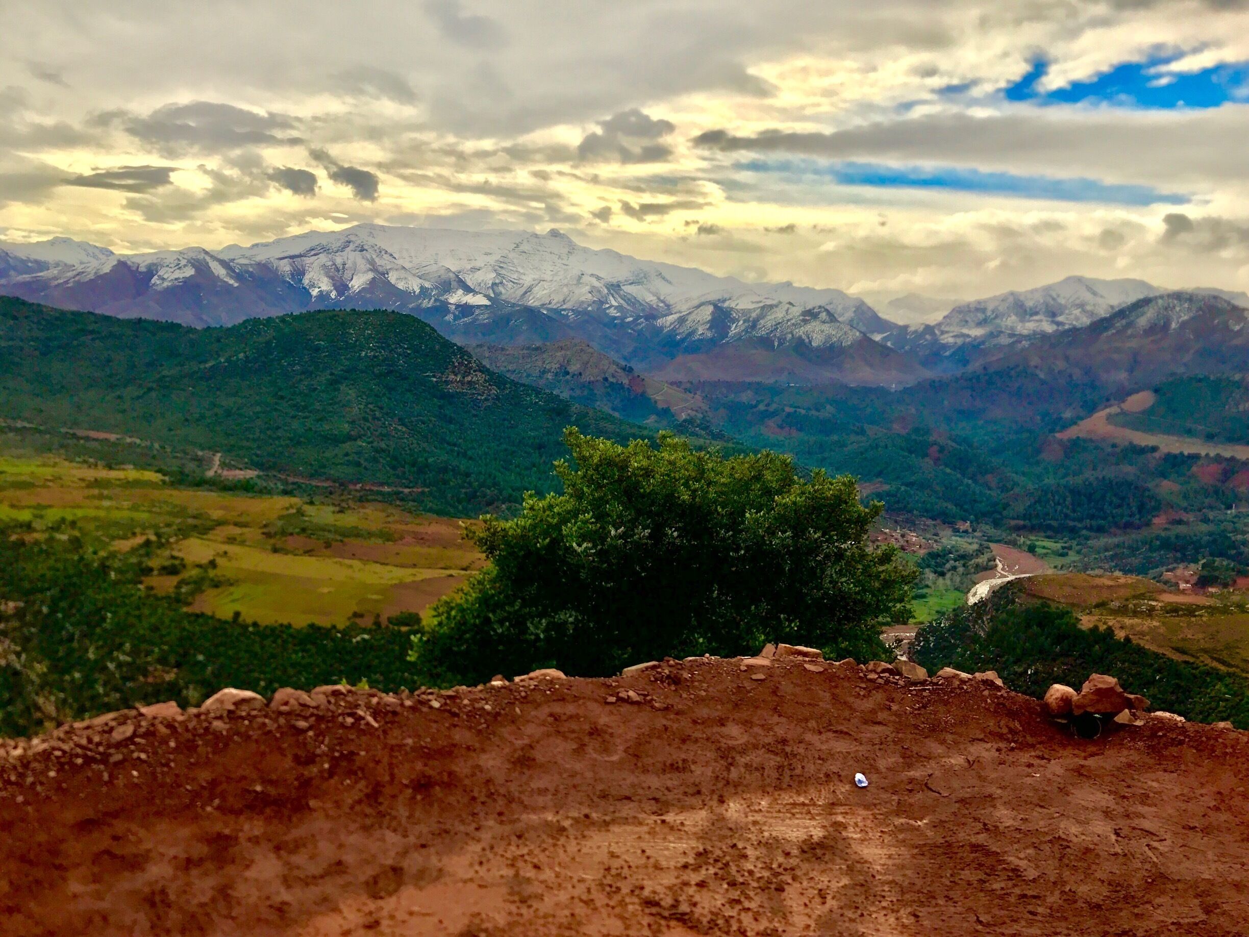 A mountainous landscape with snow-capped peaks, a valley with greenery, and a dirt path.