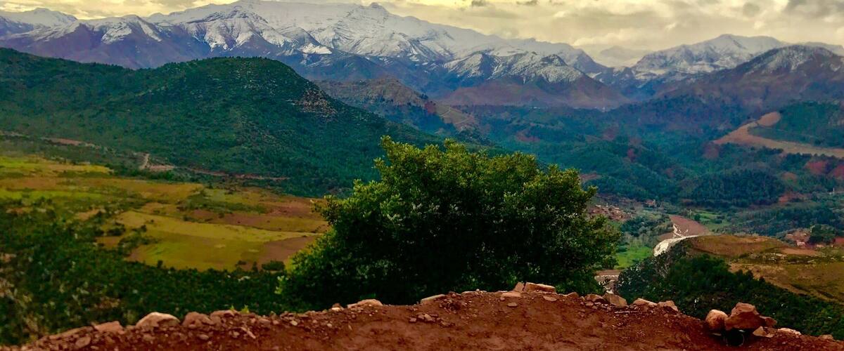 A mountainous landscape with snow-capped peaks, a valley with greenery, and a dirt path.