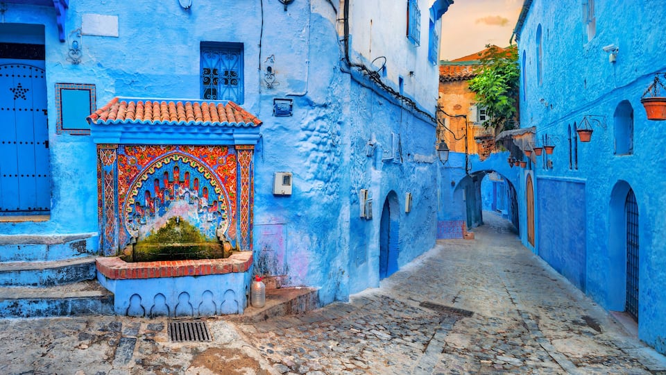 A narrow cobblestone street flanked by buildings painted in vibrant blue, with a decorative door and steps leading to a small fountain.