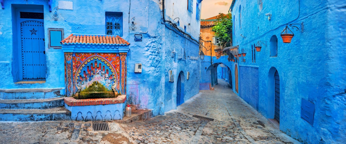 A narrow cobblestone street flanked by buildings painted in vibrant blue, with a decorative door and steps leading to a small fountain.