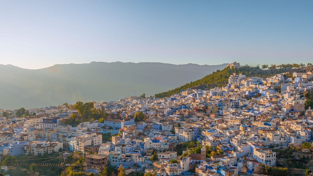 A densely packed hillside town with buildings in shades of blue and white, surrounded by mountains.