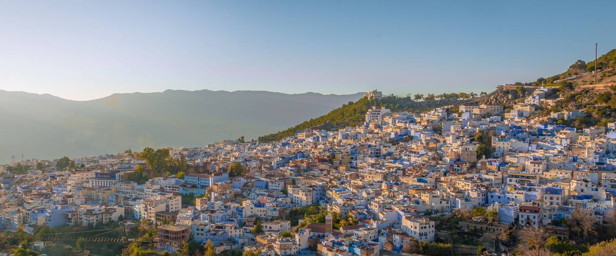 A densely packed hillside town with buildings in shades of blue and white, surrounded by mountains.