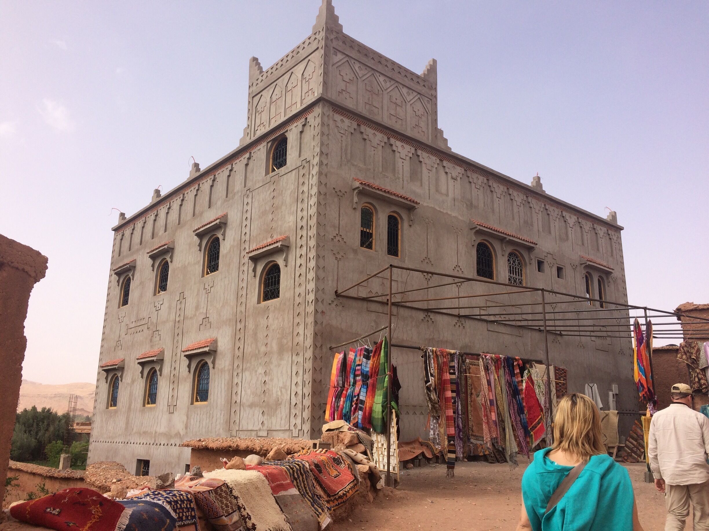 A large, historic building with arched windows and a tiled roof. In front, there is a market stall with colorful fabrics and rugs.