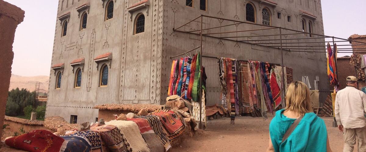 A large, historic building with arched windows and a tiled roof. In front, there is a market stall with colorful fabrics and rugs.