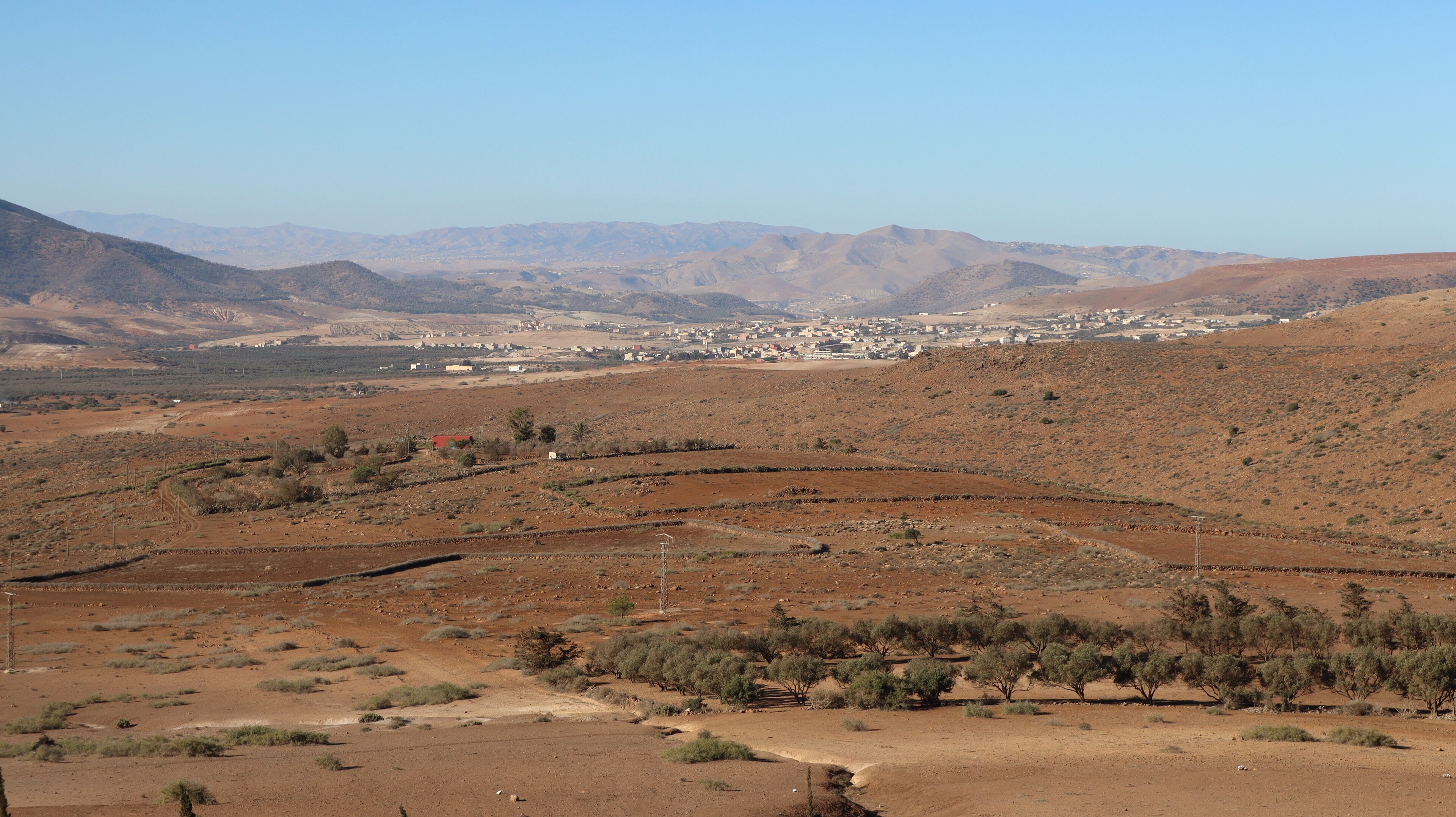 A desert landscape with sparse vegetation, a small settlement, and distant mountains.