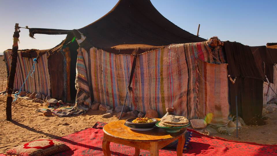 A desert tent with colorful fabrics, a wooden table with a red cloth, and a bowl of food.
