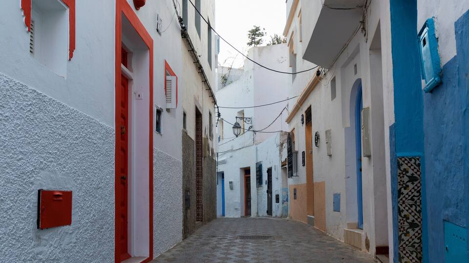 A narrow alleyway with white and blue buildings, red doors, and patterned tiles on the ground.