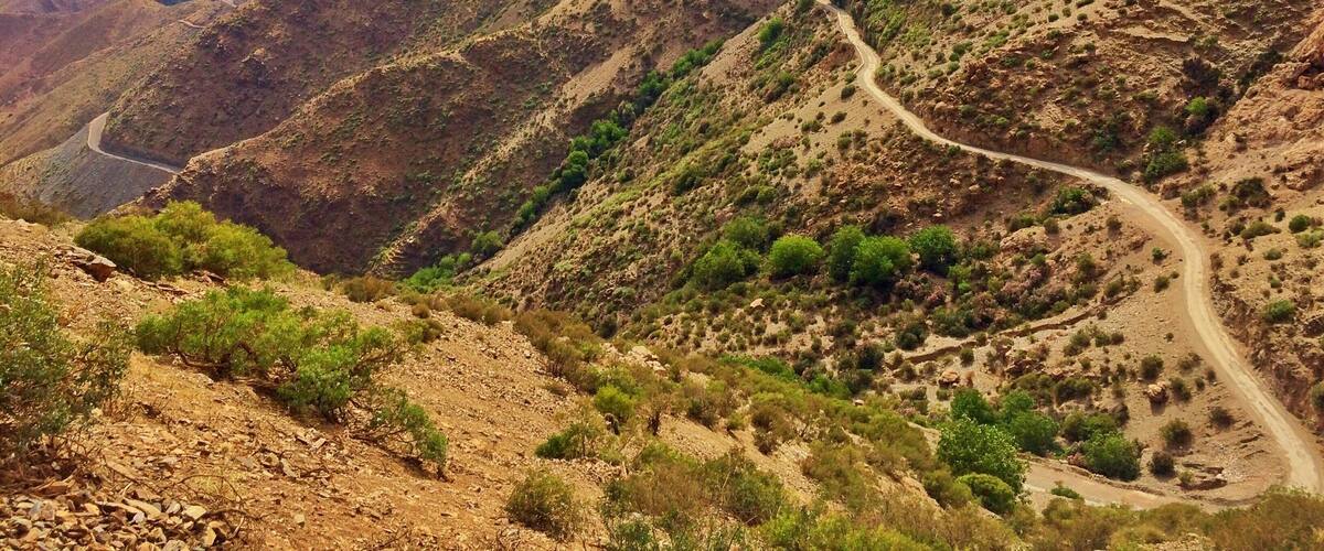 A winding mountain road through a rugged, hilly landscape with sparse vegetation and distant mountain ranges.