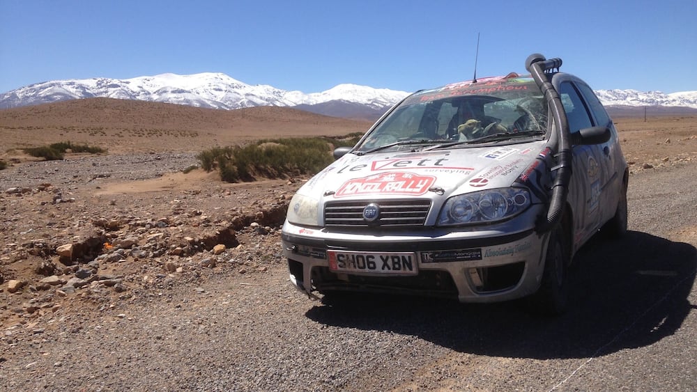 A white car with stickers on the hood parked on a rocky terrain with a mountainous background.