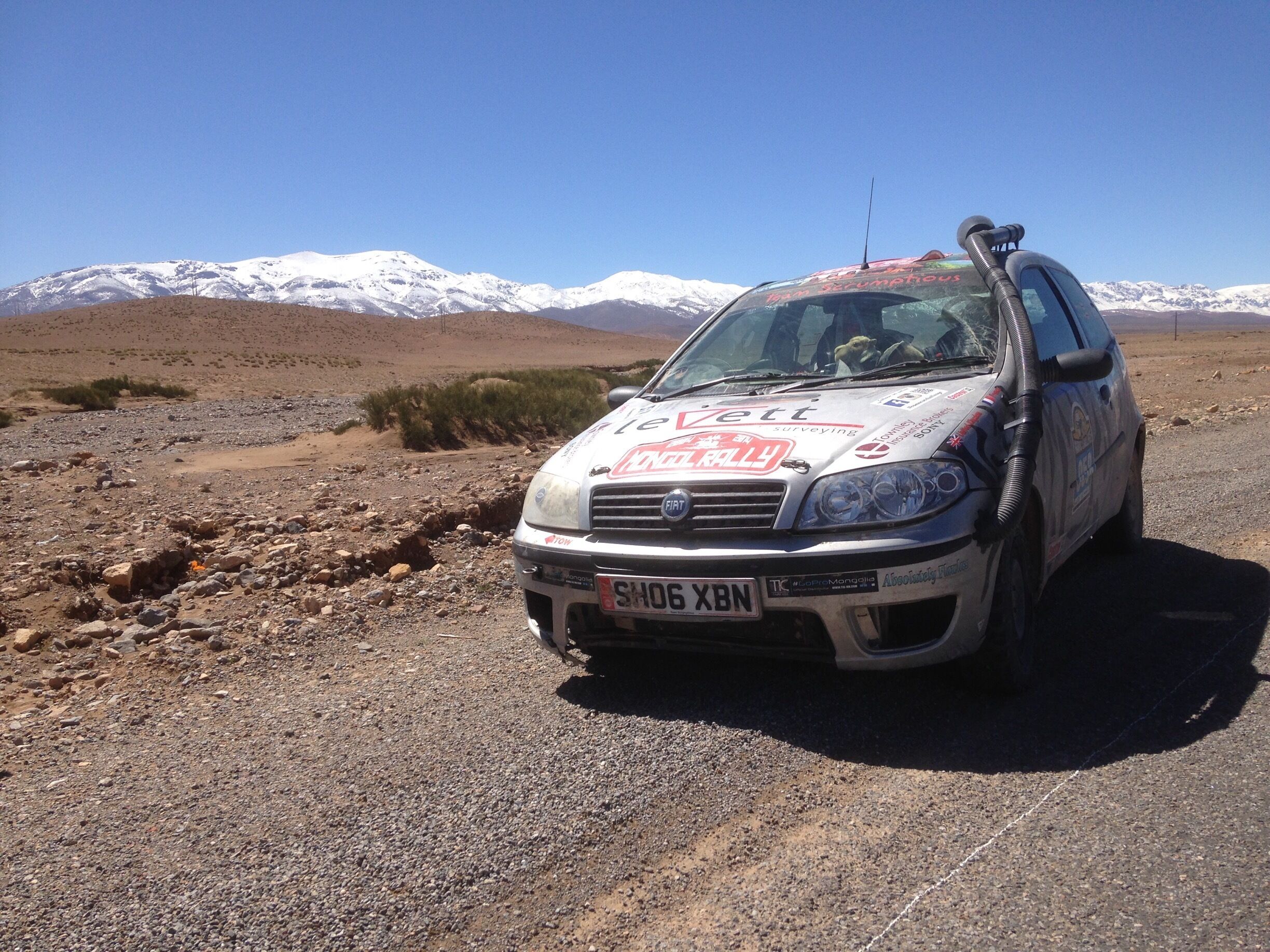 A white car with stickers on the hood parked on a rocky terrain with a mountainous background.