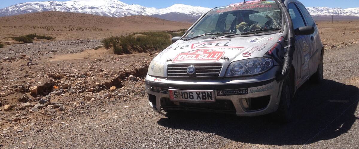 A white car with stickers on the hood parked on a rocky terrain with a mountainous background.