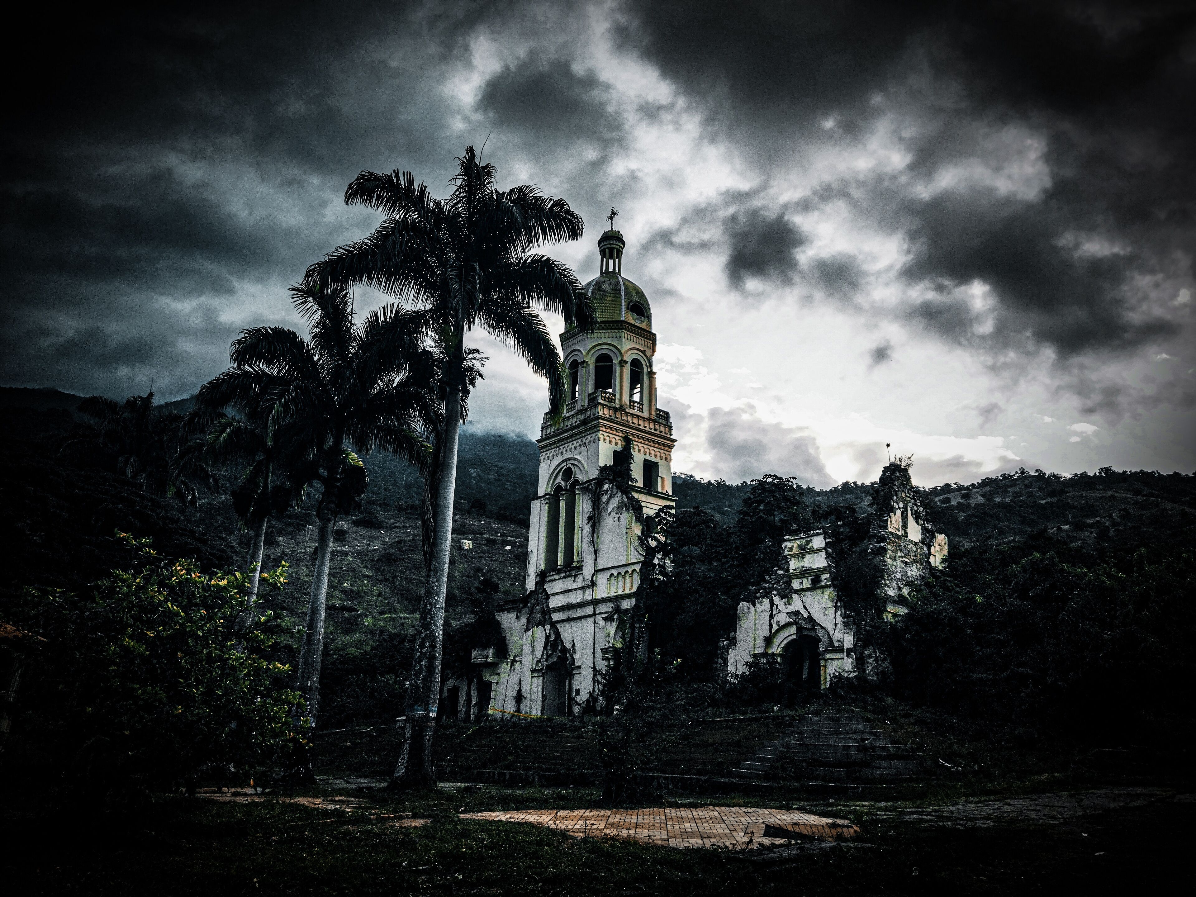 A historic church with a bell tower, surrounded by palm trees and a cloudy sky.