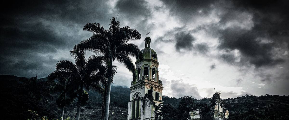 A historic church with a bell tower, surrounded by palm trees and a cloudy sky.