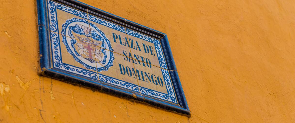 A sign with the text "PLAZA DE SANTO DOMINGO" on a yellow wall.
