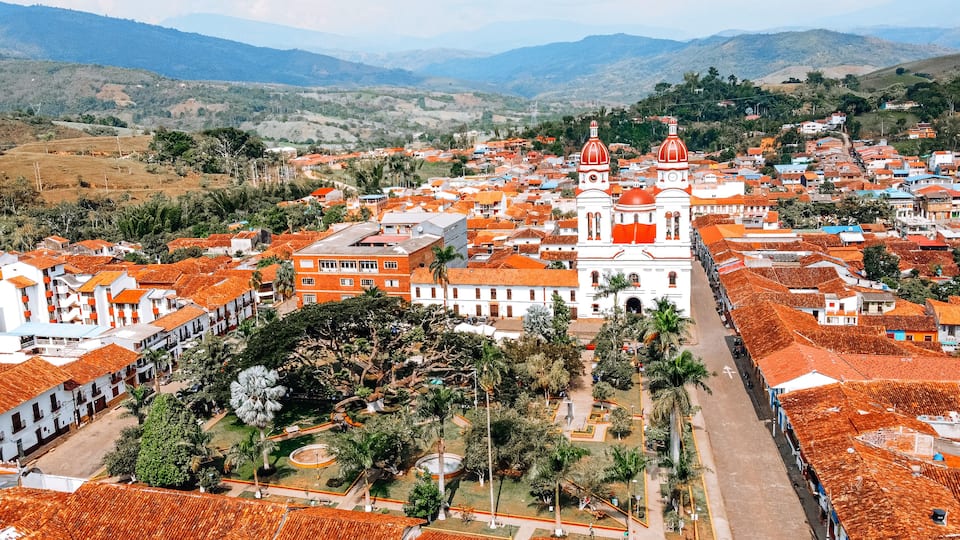A town with red-roofed buildings, a prominent white church with red domes, and surrounding green hills.
