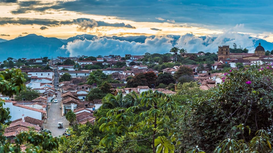 A town with terracotta-roofed buildings, a prominent church, and mountains in the background.
