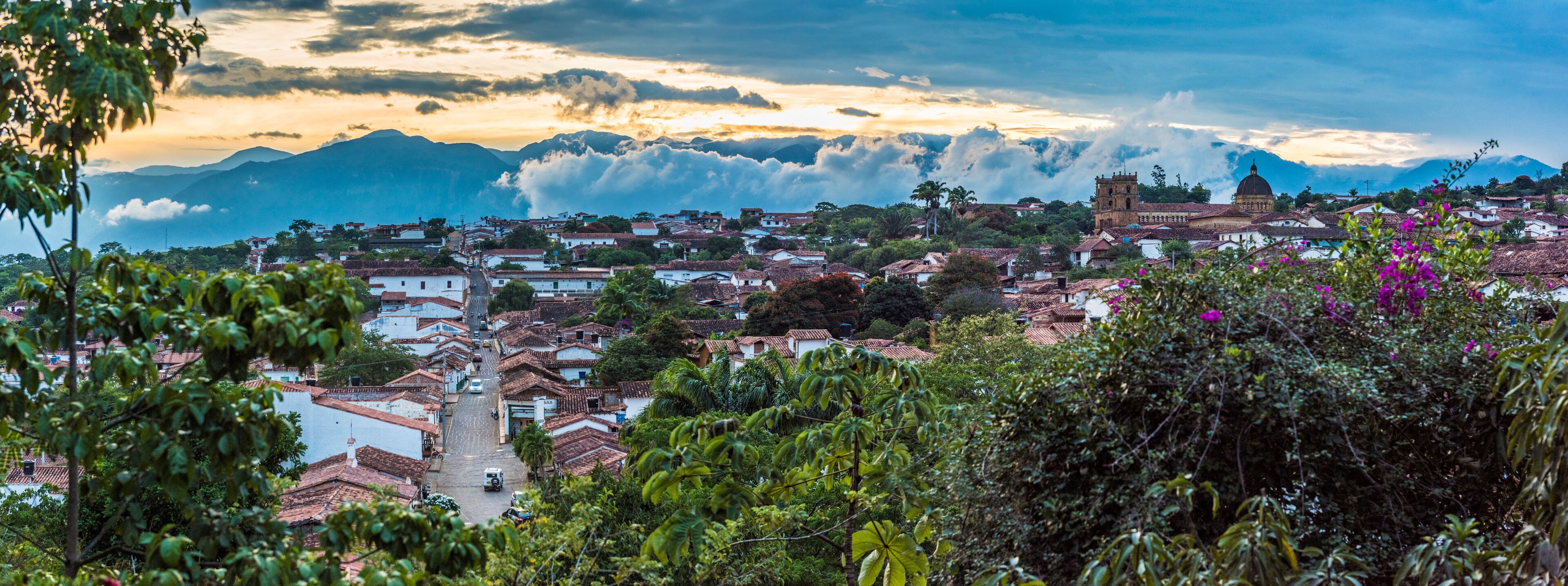A town with terracotta-roofed buildings, a prominent church, and mountains in the background.