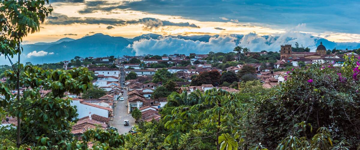 A town with terracotta-roofed buildings, a prominent church, and mountains in the background.