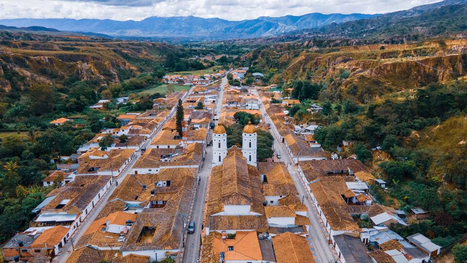 An aerial view of a town with terracotta-roofed buildings, a prominent church with two towers, and surrounding mountainous terrain.