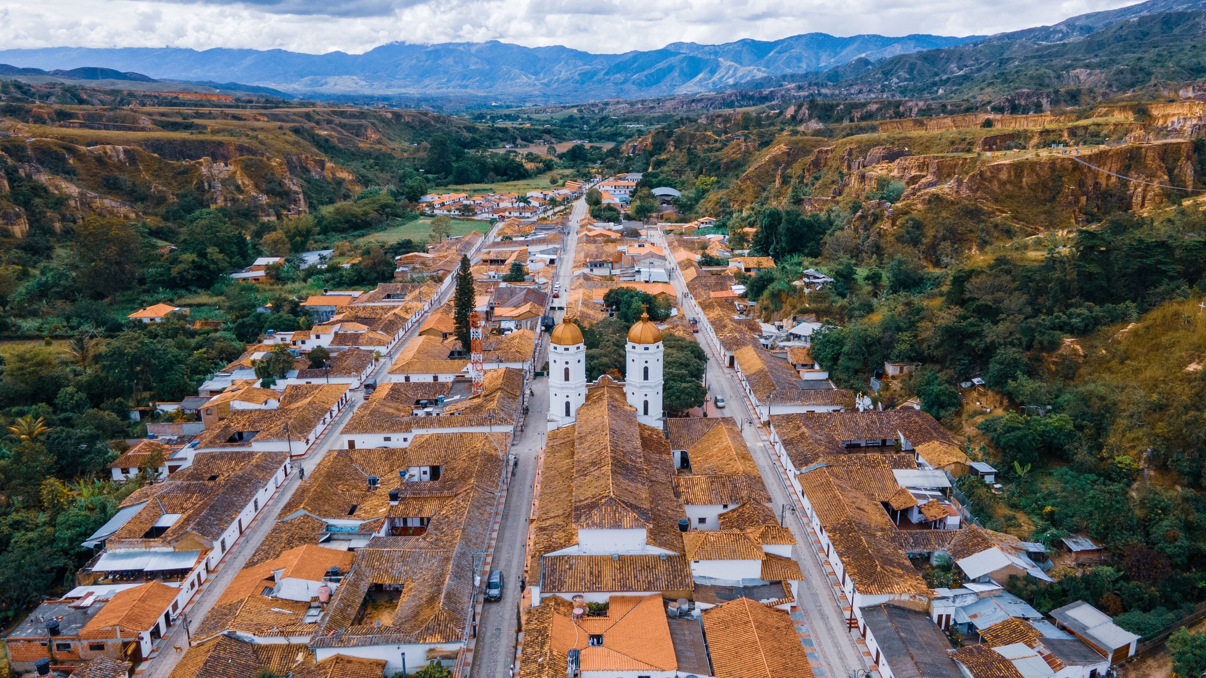 An aerial view of a town with terracotta-roofed buildings, a prominent church with two towers, and surrounding mountainous terrain.
